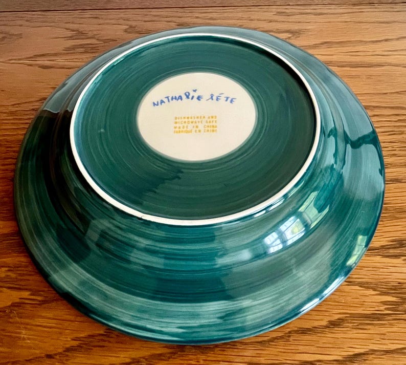 May include: A teal-colored ceramic plate with a white rim. The underside features the text "Nathalie Lete" in blue script and additional text in a smaller font. The plate has a spiral brushstroke design.