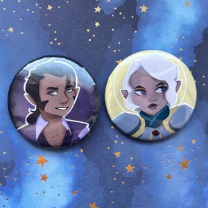 May include: Two round button pins featuring cartoon characters. The first pin features a dark-skinned character with purple hair and a purple and black outfit. The second pin features a light-skinned character with white hair and a blue and gold outfit.