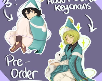 May include: Two 3-inch acrylic keychains featuring illustrated characters in teal cloaks and pointed hats. One character has dark hair and the other has blonde hair, holding a small white creature. Text reads Aggot & Coco keychains and Pre-Order.