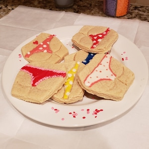 Puede incluir: Un plato con seis galletas de azúcar decoradas. Cada galleta está decorada con un diseño de glaseado de color diferente de la parte inferior de un bikini. Las galletas están en un plato blanco con chispas rosas.