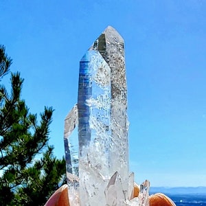 May include: A large, clear quartz crystal cluster with multiple points. The crystal is held in a hand against a blue sky background.