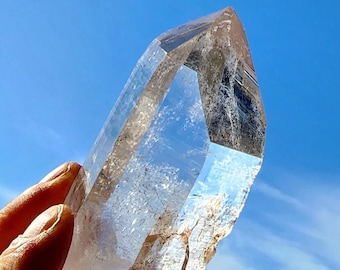 May include: A large, clear quartz crystal with a double termination. The crystal is held in a hand against a blue sky background.