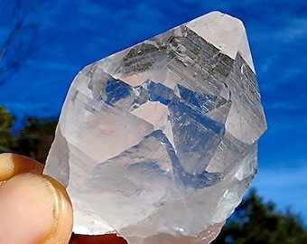 May include: A clear quartz crystal with a pointed tip and multiple facets. The crystal is held in a hand against a blue sky background.