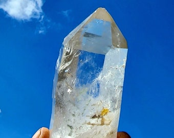 May include: A large, clear quartz crystal with a pointed tip. The crystal is held in a hand against a blue sky with white clouds.