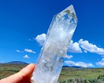 May include: A large, clear quartz crystal point with a natural, rough texture. The crystal is held in a hand against a backdrop of a blue sky with white clouds and a green grassy field.