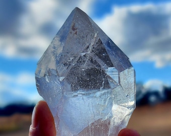 May include: A large, clear quartz crystal with a pointed tip and many facets. The crystal is being held in a hand against a blue sky with white clouds.