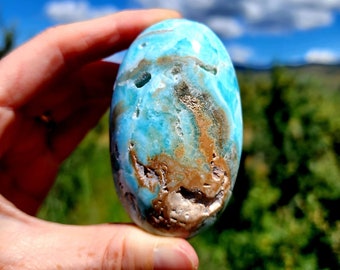 May include: A smooth, oval-shaped blue and brown gemstone with a swirling pattern. The gemstone is being held in a hand.