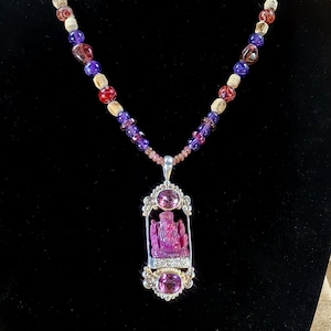 May include: A beaded necklace featuring a pendant with a carved pink figure. The necklace has a mix of purple, red, white, and brown beads. The pendant is framed with silver-colored metal and has two oval-shaped pink stones.