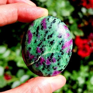 May include: A green and pink gemstone with black flecks. The stone is smooth and oval shaped.
