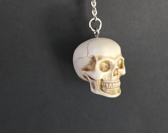 May include: A skull-shaped keychain with a silver chain and ring. The skull is off-white with detailed facial features, including teeth and eye sockets. The keychain is suspended against a dark gray background.