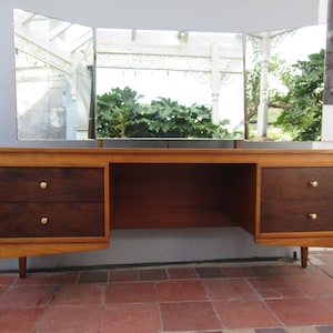 May include: A wooden vanity table featuring a light brown frame with two sets of two dark brown drawers on either side. Each drawer has a small round knob. A tri-fold mirror with three rectangular glass panels is attached to the top surface.