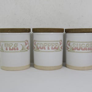 May include: Three white ceramic canisters with wooden lids labelled "TEA", "COFFEE", and "SUGAR". Each canister has a decorative floral design in pink and green. The canisters are cylindrical and have a light beige base.