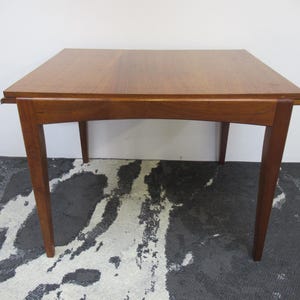 May include: A square wooden table with a warm brown finish. The table has a simple design with four angled legs and a smooth tabletop. The table measures approximately 76 cm wide and 76 cm long.