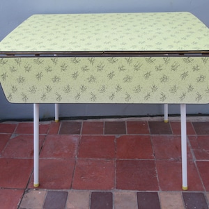May include: A rectangular table with a light yellow laminate top and drop-down sides, featuring a dark green abstract botanical pattern. The table is supported by four slender, straight white metal legs with yellow caps at the base of each leg.