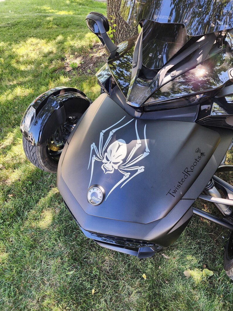 A black three-wheeled motorcycle with a skull and spider design on the front, parked on a grassy area.