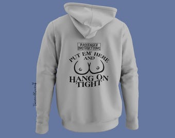 Puede incluir: Una sudadera con capucha de color gris claro que presenta un gráfico negro en la parte posterior. El texto dice PASSENGER INSTRUCTIONS, PUT EM' HERE AND HANG ON TIGHT, junto a un dibujo lineal de pechos. El nombre de la marca TwistedRoots está impreso verticalmente.