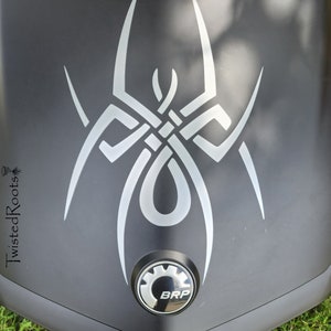 May include: A matte black surface featuring a large, stylized silver spider decal with intricate, interwoven lines. Below the spider design is a circular emblem with a gear-like border and the letters BRP. The text TwistedRoots is visible on the left.