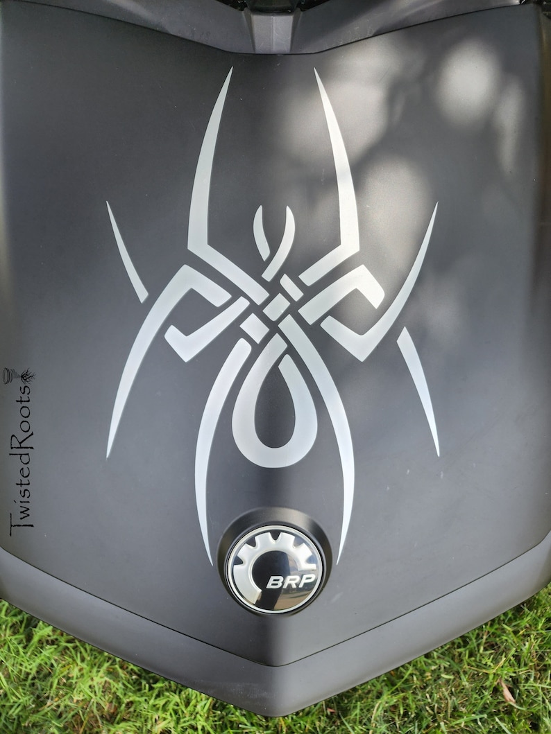 May include: A matte black surface featuring a large, stylized silver spider decal with intricate, interwoven lines. Below the spider design is a circular emblem with a gear-like border and the letters BRP. The text TwistedRoots is visible on the left.