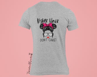 Puede incluir: Una camiseta gris jaspeado de manga corta con un gráfico de una persona con un moño desordenado, un lazo rosa brillante y gafas. El texto dice "Ryker Hair DON'T CARE!" en negro, con labios rojos. El nombre de la marca "TwistedRoots" está impreso verticalmente.