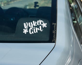 May include: A white vinyl decal applied to a car window featuring the text Ryker Girl in a cursive script font, flanked by two simple flower icons. A smaller vertical text element reads TwistedRoots along the right edge of the window glass.