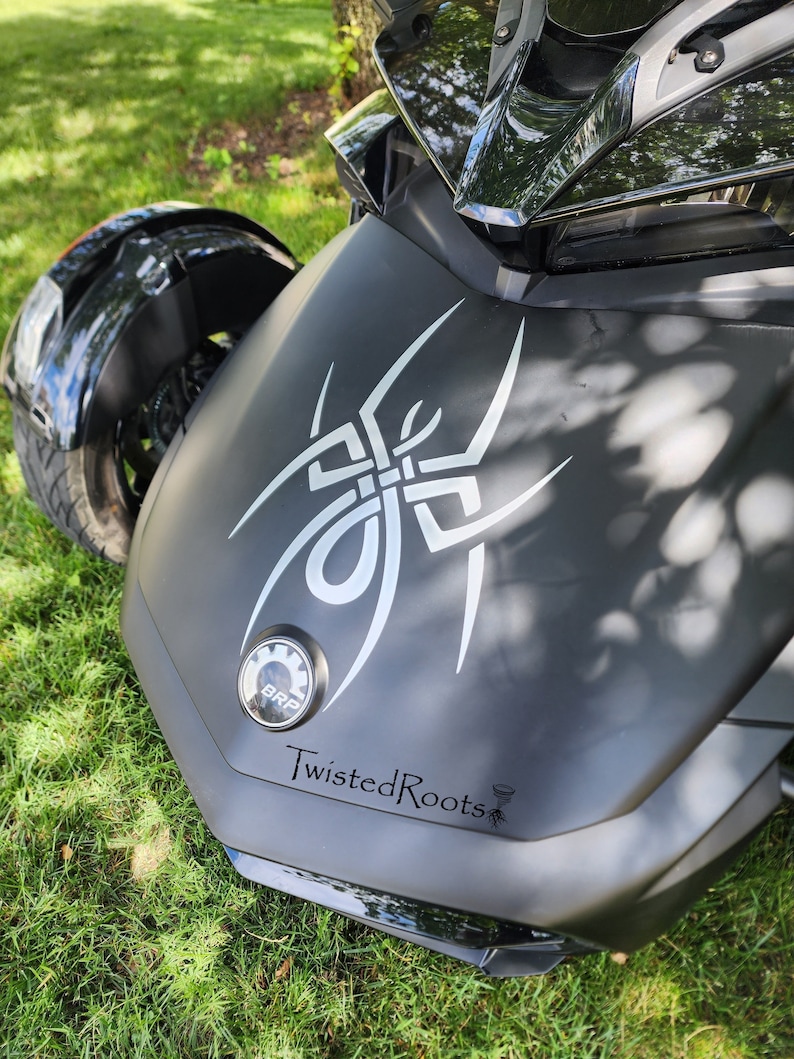 May include: A matte black three-wheeled motorcycle featuring a large white stylized spider decal on the front hood. A circular BRP emblem is positioned below the spider, and the text TwistedRoots is printed in black lettering near the bottom.