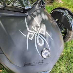May include: A matte black three-wheeled motorcycle hood featuring a large white tribal spider decal, a circular BRP emblem, and the text TwistedRoots with a small tornado and root graphic printed in black on the lower section of the front panel.
