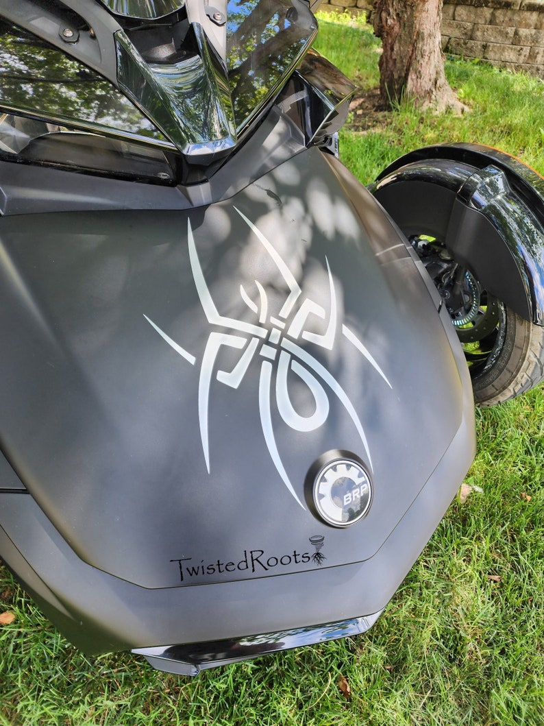 May include: A matte black three-wheeled motorcycle hood featuring a large white tribal spider decal, a circular BRP emblem, and the text TwistedRoots with a small tornado and root graphic printed in black on the lower section of the front panel.