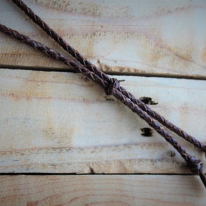 May include: A brown leather cord with a braided design and a brass ring at the end. The cord is lying on a wooden surface.