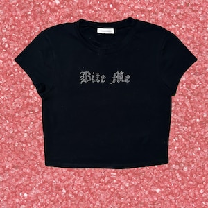 May include: A black short-sleeved crop top featuring the text "Bite Me" written in silver-toned rhinestones across the chest. The garment has a round neckline and a straight hemline, with a small white brand label visible at the inner collar.