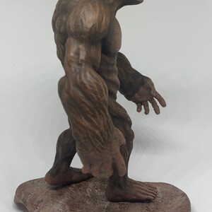 Może przedstawiać: Brązowa, rzeźbiona figurka Bigfoota stojąca na skalnym cokole. Figurka jest szczeg&oacute;łowo wykonana z futra i definicją mięśni.