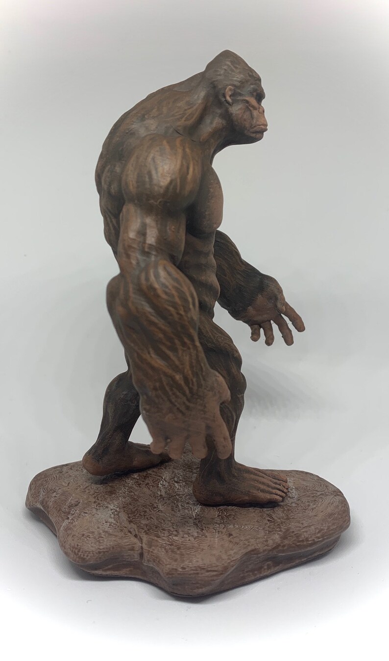 Może przedstawiać: Brązowa, rzeźbiona figurka Bigfoota stojąca na skalnym cokole. Figurka jest szczeg&oacute;łowo wykonana z futra i definicją mięśni.