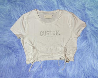 May include: White crop top with a drawstring detail at the bottom. The top has the word "CUSTOM" written in rhinestones.