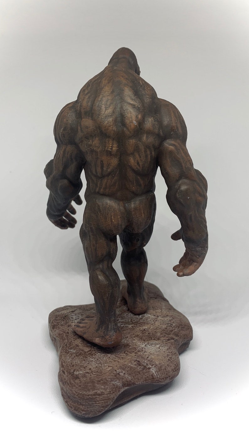 Może przedstawiać: Brązowa, szczeg&oacute;łowa, rzeźbiona figurka muskularnej istoty humanoidalnej stojącej na skalnej podstawie. Figurka jest ustawiona w pozycji chodzenia, z jedną nogą do przodu i drugą do tyłu.