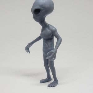 Puede incluir: Una figurita de extraterrestre gris con una cabeza grande y extremidades largas y delgadas. La figurita est&aacute; de pie con los brazos extendidos y las piernas ligeramente dobladas.