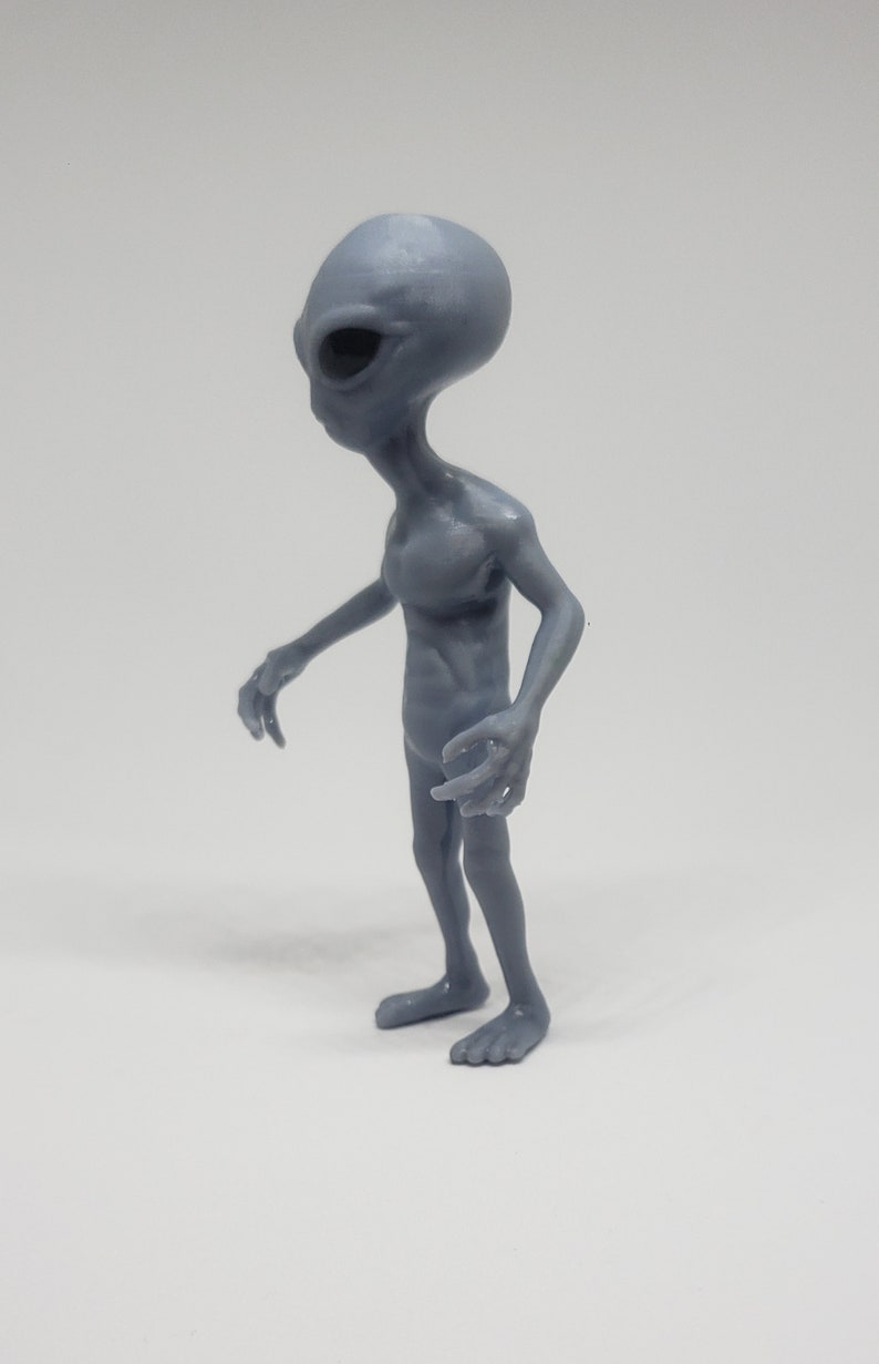 Puede incluir: Una figurita de extraterrestre gris con una cabeza grande y extremidades largas y delgadas. La figurita est&aacute; de pie con los brazos extendidos y las piernas ligeramente dobladas.