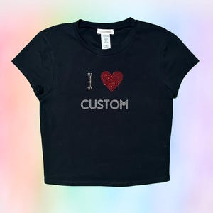 Op de afbeelding: Zwart getailleerd T-shirt met een rood hart van strass en de tekst "I &hearts; CUSTOM" in zilveren strass.