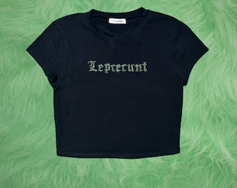 Op de afbeelding: Zwarte crop top met het woord "Leprecunt" in groene strass steentjes. De top heeft korte mouwen en een ronde hals. De achtergrond is een fuzzy, heldergroen oppervlak.