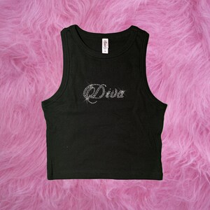 May include: Black tank top with the word "Diva" spelled out in rhinestones. The top is sleeveless and has a scoop neck. The tank top is on a pink, fluffy surface.