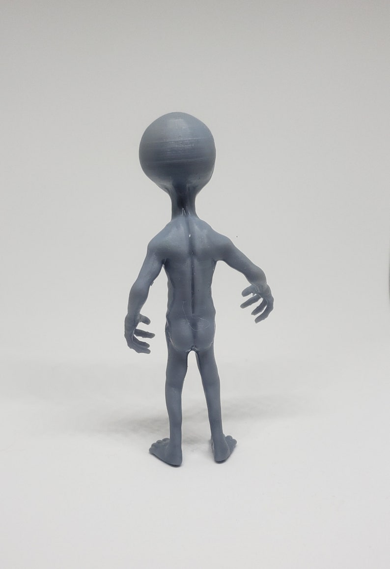 Puede incluir: Una figurita de extraterrestre gris de pie con los brazos extendidos. La figurita est&aacute; hecha de pl&aacute;stico y tiene un acabado liso y mate.