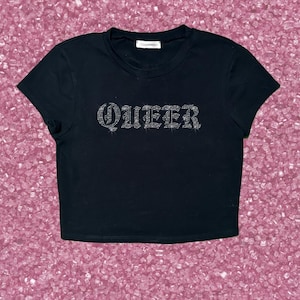 May include: Black crop top with the word "QUEER" spelled out in rhinestones. The shirt has a crew neck and short sleeves. The top is displayed on a pink background.