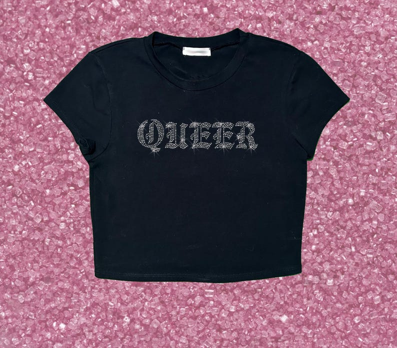 May include: Black crop top with the word "QUEER" spelled out in rhinestones. The shirt has a crew neck and short sleeves. The top is displayed on a pink background.