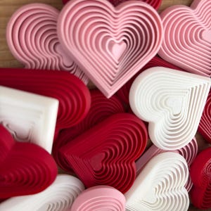 May include: A collection of heart-shaped objects in various shades of red, pink, and white. The hearts are layered, creating a three-dimensional effect. The objects are likely cookie cutters or decorative items, perfect for Valentine's Day or other celebrations.