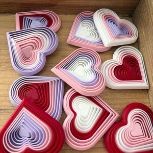 May include: A collection of layered heart-shaped decorations in various colors, including red, pink, white, and lavender. The hearts are nested within each other, creating a 3D effect. They are arranged on a wooden surface.