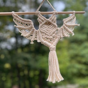 May include: A cream-colored macrame wall hanging shaped like a bat, crafted from cotton cord with intricate knotting patterns. The bat figure is suspended from a smooth, horizontal wooden dowel and features a long, fringed tassel at the bottom.