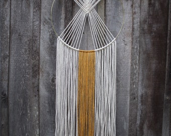 May include: A wall hanging featuring a circular gold metal hoop with off-white cotton cord knotted in a geometric pattern at the top. Long, vertical strands of off-white and mustard yellow cord hang from the bottom half of the hoop.