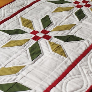 Peut inclure: Un chemin de table matelassé blanc avec un motif géométrique de diamants verts, dorés et rouges. Le chemin de table a une bordure rouge avec un bord festonné.