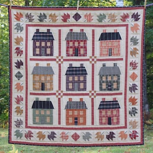 Peut inclure: Une courtepointe avec un motif de patchwork représentant neuf maisons de différentes couleurs et motifs. Les maisons sont entourées d'une bordure de feuilles rouges, blanches et brunes.