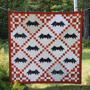 May include: A Halloween-themed quilt featuring a repeating pattern of black bat silhouettes on a white background. The quilt has a border of orange and brown squares, with a matching border. The quilt is hanging outside on a clothesline.