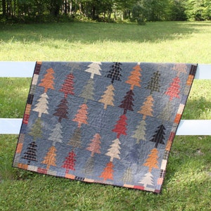 May include: A handmade quilt featuring a pattern of colorful trees on a blue-gray background. The trees are in various shades of brown, red, and green. The quilt is draped over a white fence, with a green grassy background.