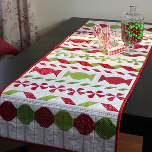 May include: A white, red, and green quilted table runner with a candy cane and peppermint design. The runner has a scalloped edge and is decorated with red and green hexagons.